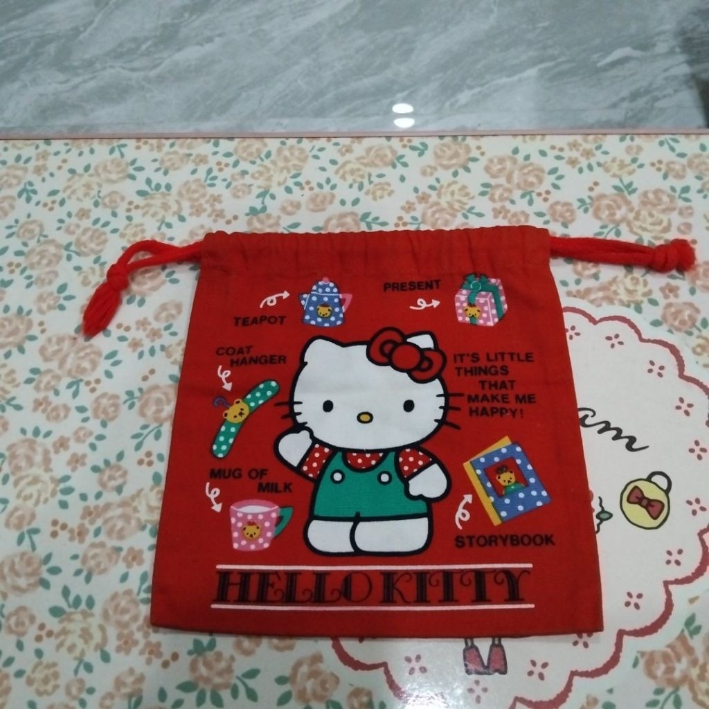กระเป๋าผ้าหูรูด Hello Kitty สีแดงในภาพเป็นสินค้าวินเทจจาก Sanrio ลวดลายบนกระเป๋ามีคิตตี้สวมชุดเอี๊ยมสีเขียว